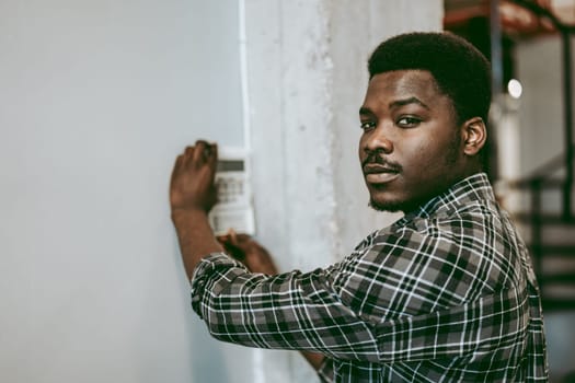A man wearing a plaid shirt is focused on adjusting a device mounted on the wall. He is in a modern industrial setting, illuminated by natural light.