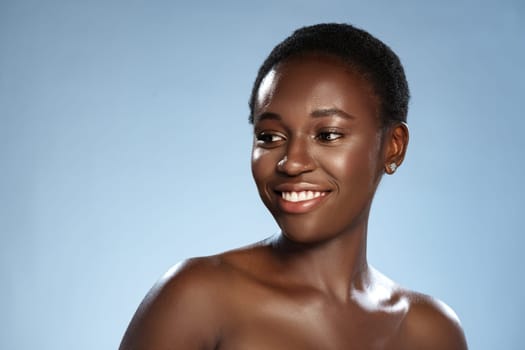 A woman with short hair smiles confidently while posing against a soft light blue background. The lighting highlights her glowing skin and joyful expression, embodying beauty.