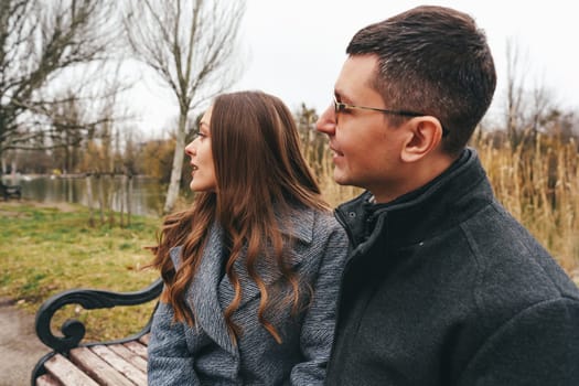 A couple sits on a bench by a serene lake in a park, wrapped in warm clothing. They gaze in opposite directions, enjoying the tranquil autumn atmosphere and each other's company.