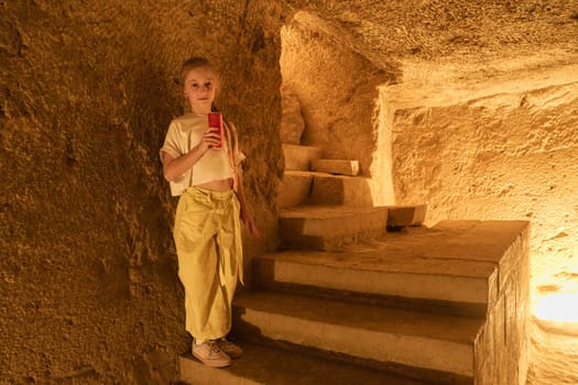 A young girl stands beside stone steps in a cavernous space, surrounded by warm, golden light. She is focused on her phone, capturing the moment in the underground cave.