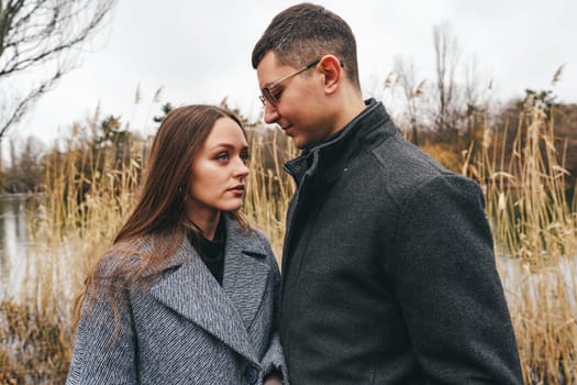 A couple is facing each other near a lake, deeply engaged in conversation. They are surrounded by tall grass and the overcast sky adds a moody atmosphere to the setting.