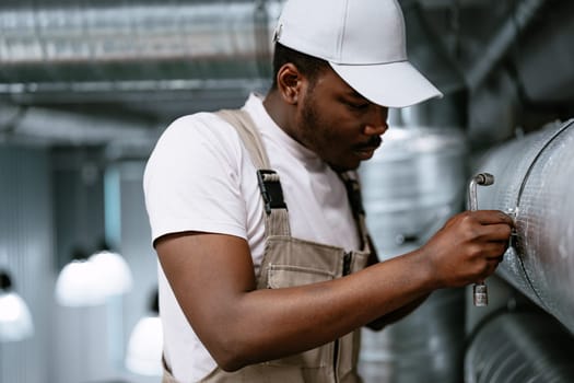 A diligent worker focuses on fixing ductwork in an industrial space, wearing safety attire. The bright environment indicates daytime operations with metallic surfaces surrounding him.