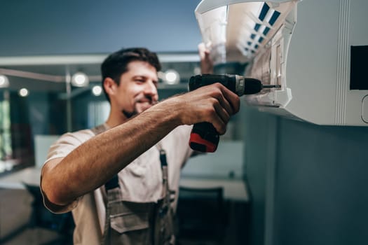 A skilled technician works on an air conditioning unit inside a contemporary office space. He uses a power drill to conduct necessary repairs, smiling as he focuses on the task.