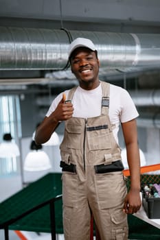 A cheerful young technician wearing beige overalls stands confidently in a well-lit, modern workspace, giving a thumbs up. The atmosphere exudes professionalism.