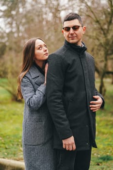 A woman in a gray coat affectionately leans against a man dressed in a black jacket, both smiling while surrounded by green trees in a park setting.