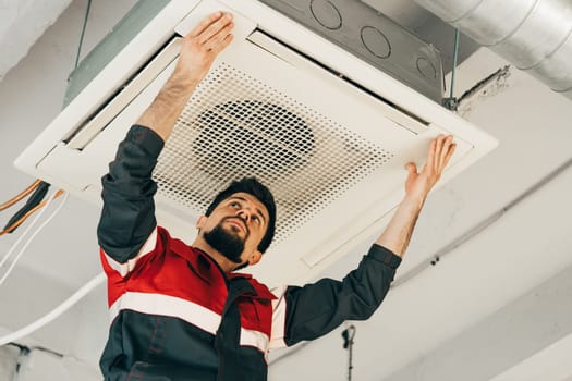 A technician is focused on installing a ceiling-mounted air conditioning unit in a contemporary office setup. The workspace is well-lit and features exposed ductwork.