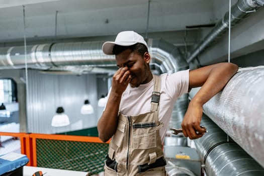 A young man, dressed in coveralls and a cap, appears tired as he leans against a metal pipe in a well-lit industrial space. He is likely taking a moment during repairs.