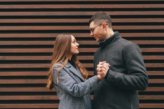 A young couple shares a tender moment, gazing into each other's eyes while holding hands. They are positioned in front of a stylish wooden wall, suggesting a modern atmosphere.