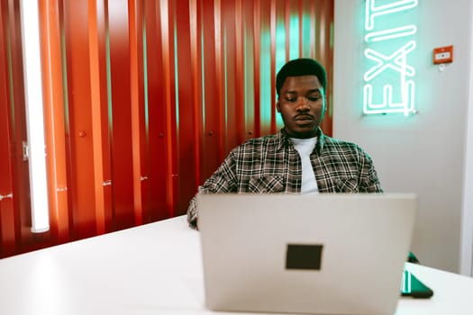 A focused young man sits at a minimalistic table, using his laptop in a lively coworking space with bright orange walls and glowing green exit signage.