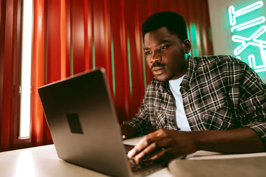 A man with dark skin sits at a table in a stylish workspace, intently typing on his laptop. Colorful neon light accents enhance the modern atmosphere as he works late.