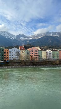 Innsbruck, Austria - March 28, 2025: Charming pastel-colored buildings line a riverbank Inn against a backdrop of snow-capped Alpine mountains and clear blue sky, providing a picturesque view of a serene and vibrant European town.