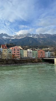 Innsbruck, Austria - March 28, 2025: Riverside houses painted in vibrant colors, with a scenic backdrop of snow-capped mountains and a serene river Inn in the foreground, creating a picturesque and tranquil natural urban scene.