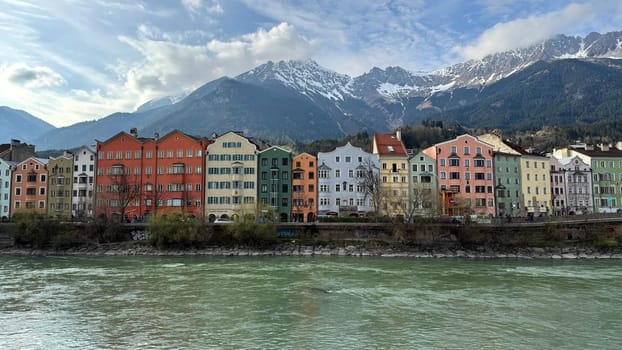 Innsbruck, Austria - March 28, 2025: Vibrant row of colorful houses along the Inn River in Innsbruck, Austria, with snow-capped mountains in the background. A picturesque European cityscape in spring light, blending traditional architecture and natural alpine beauty.