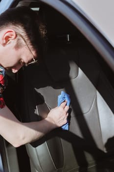 Portrait of one young Caucasian brunette man who is leaning over cleaning leather seat inside car interior with cloth and cleaning agent on sunny spring day at car wash, top view from side very close up with selective focus. Concept car wash, working people.