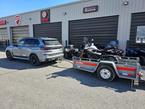 Ottawa, ON Canada - 03.15.2024: Silver BMW X5 SUV Towing a Yamaha Tracer 9 GT Motorcycle on a Trailer Outside Dealership, Automotive Transport Concept.