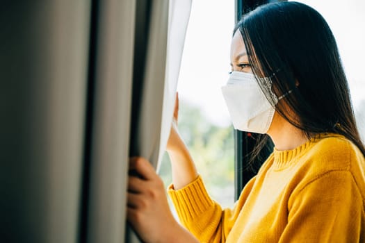 A young Asian woman in a medical mask looks through the window feeling upset and distressed due to COVID-19. Reflecting fear isolation and pandemic-related anxiety.