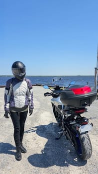 A female motorcyclist in full gear stands beside her motorcycle on a scenic lakeside road. The sunny day and beautiful landscape evoke a sense of freedom and adventure.