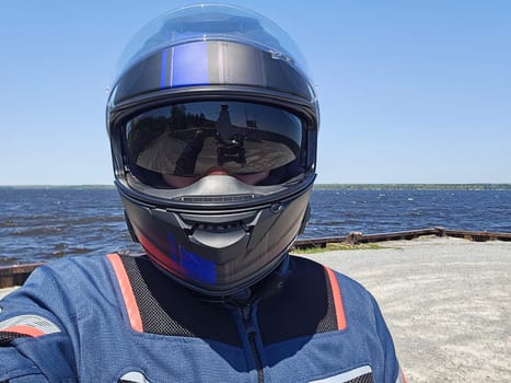 A motorcycle rider takes a selfie wearing a dark helmet with a reflective visor. The scenic lake and sunny day create a beautiful backdrop for the rider in a blue and black jacket.