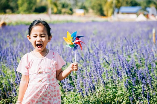 Cheerful little girl holds a toy pinwheel in a flower garden. Spring's joy and the spinning pinwheel symbolize childhood happiness fun and freedom in the sunny embrace of nature.