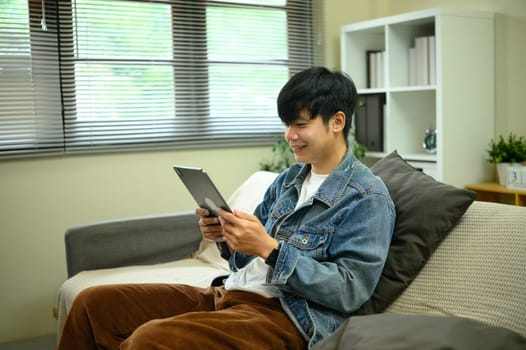 Happy young asian man wearing denim jacket using digital tablet in living room.