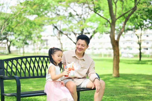 Father and daughter sit on a park bench enjoying a bubble toy on a sunny day.