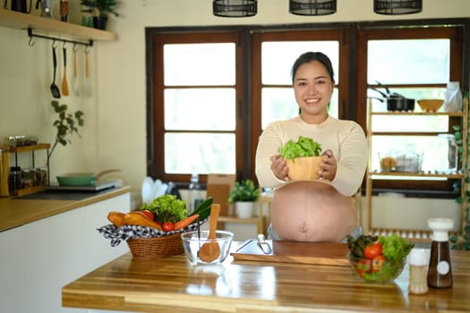 Asian pregnant woman standing in a bright kitchen, presenting a bowl of salad.