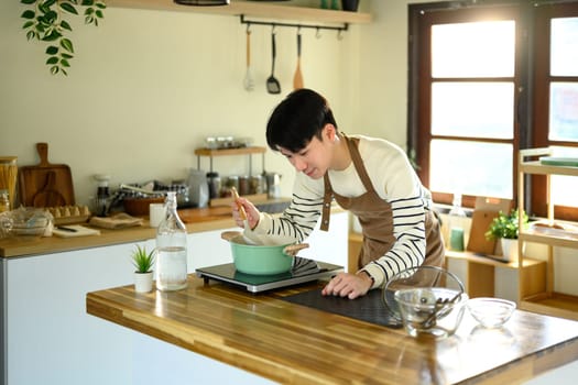 Happy Asian man preparing a meal on a modern stovetop.