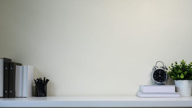 Minimal desk with books, alarm clock, and green potted plant against a blank beige wall.