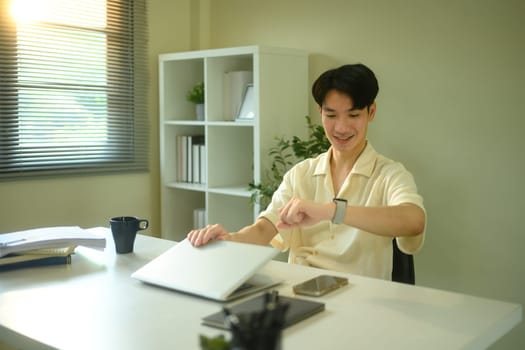 Smiling young man checking the time on his smartwatch and closing a laptop.
