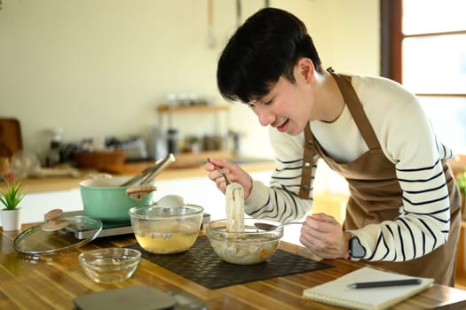 Smiling young man trying freshly cooked noodles on kitchen counter.