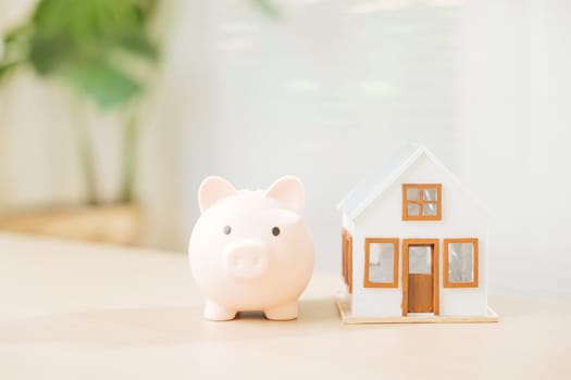 A piggy bank and white house model sit on a table, symbolizing saving money for property investment. Explore real estate opportunities, dream home buying, and smart mortgage planning.
