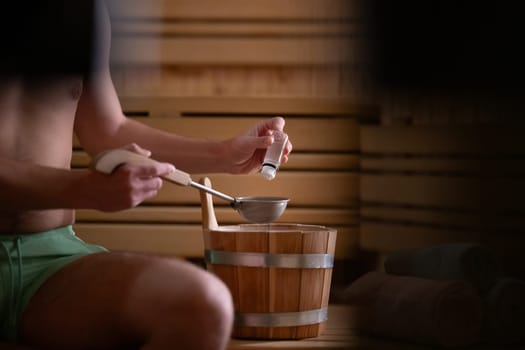 Close up of a mans hand holding essential oils inside a traditional wooden sauna with bucket and ladle.