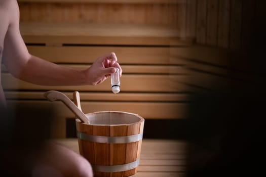 Close up of a mans hand holding essential oils inside a traditional wooden sauna with bucket and ladle.