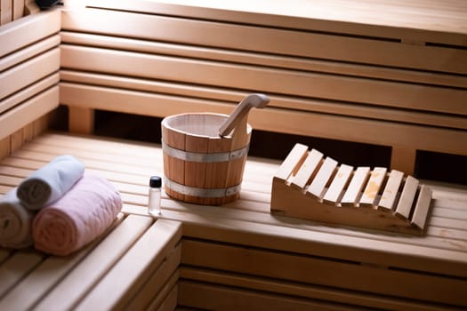 Detailed close up of sauna bucket with ladle and essential oils next to rolled towels on wooden bench inside sauna.