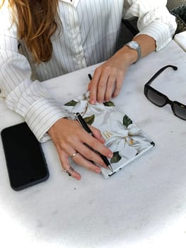 A person with long hair is writing with a pen in a floral notebook, seated at a cafe table under sunlight.