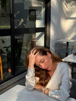 A woman with long hair rests her head in her hand while sitting at a cafe table, lost in thought, illuminated by soft afternoon light.