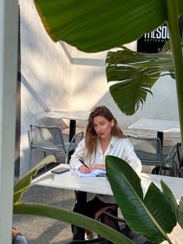 A woman sits at a table in an outdoor cafe, writing in a notebook, with tropical plants around her. The sun shines softly.