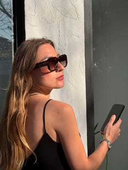 A woman with long hair stands beside a window, holding a smartphone while enjoying the sunlight on her face.