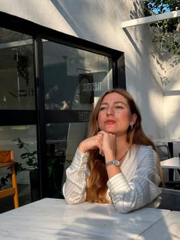 A young woman relaxes at a light-filled cafe, gazing thoughtfully as sunlight enhances her serene expression.