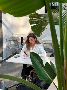 A young woman focuses on her studies at an outdoor cafe, surrounded by plants and enjoying a sunny afternoon.