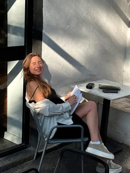 A young woman relaxes outdoors at a cafe, smiling and writing in her notebook while basking in the warm afternoon sun.