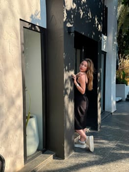 A fashionable woman stands elegantly against a sleek wall, showcasing contemporary design and bright sunlight in a lively city.