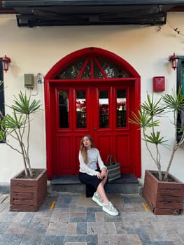A woman relaxes on steps outside a cafe with a striking red door and stylish potted plants. The urban setting adds to the charm.