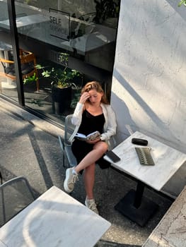 A young woman sits at a table in a stylish cafe, reading a book while enjoying the natural light streaming in.