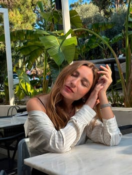 A young woman sits at a cafe table, lost in thought, surrounded by vibrant plants and greenery on a sunny day.