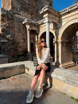 A young woman enjoys a sunny day at historical ruins, seated comfortably on a stone ledge, soaking in the surroundings.