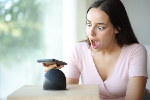 Amazed woman pairing smart speaker with mobile phone sitting on a couch at home