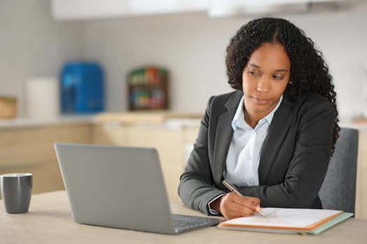 Black executive working online taking notes in a notebook and checking laptop at home
