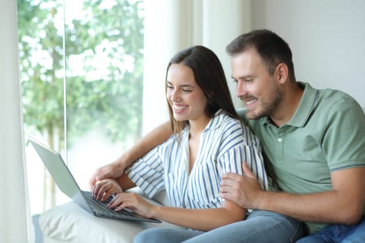 Happy couple using a laptop together sitting in a couch in a house interior