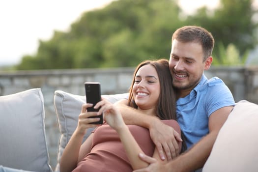 Happy couple smiling checking mobile phone sitting in a couch in a terrace at home
